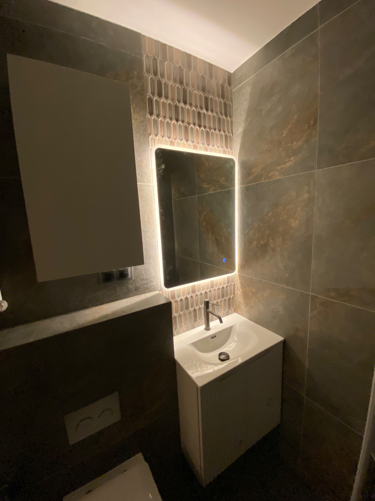 Nowoczesna łazienka z podświetlanym lustrem, umywalką z szafką i mozaiką w kształcie rombów. Ciemne płytki imitujące kamień na ścianach i podłodze.