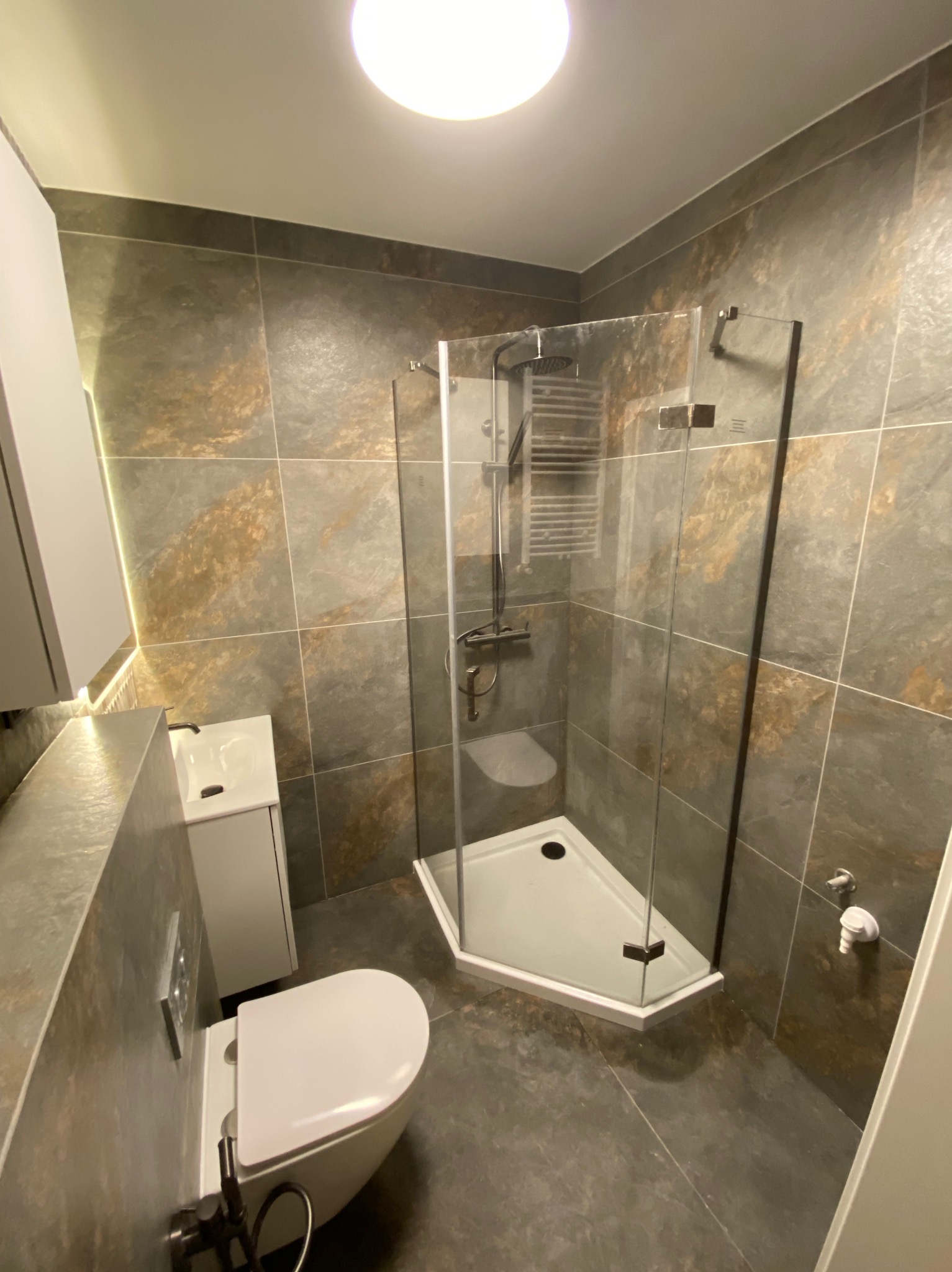 Nowoczesna łazienka z kabiną prysznicową narożną, umywalką z szafką i podwieszaną toaletą. Płytki w odcieniach szarości i brązu z delikatnym wzorem. Oświetlenie sufitowe.