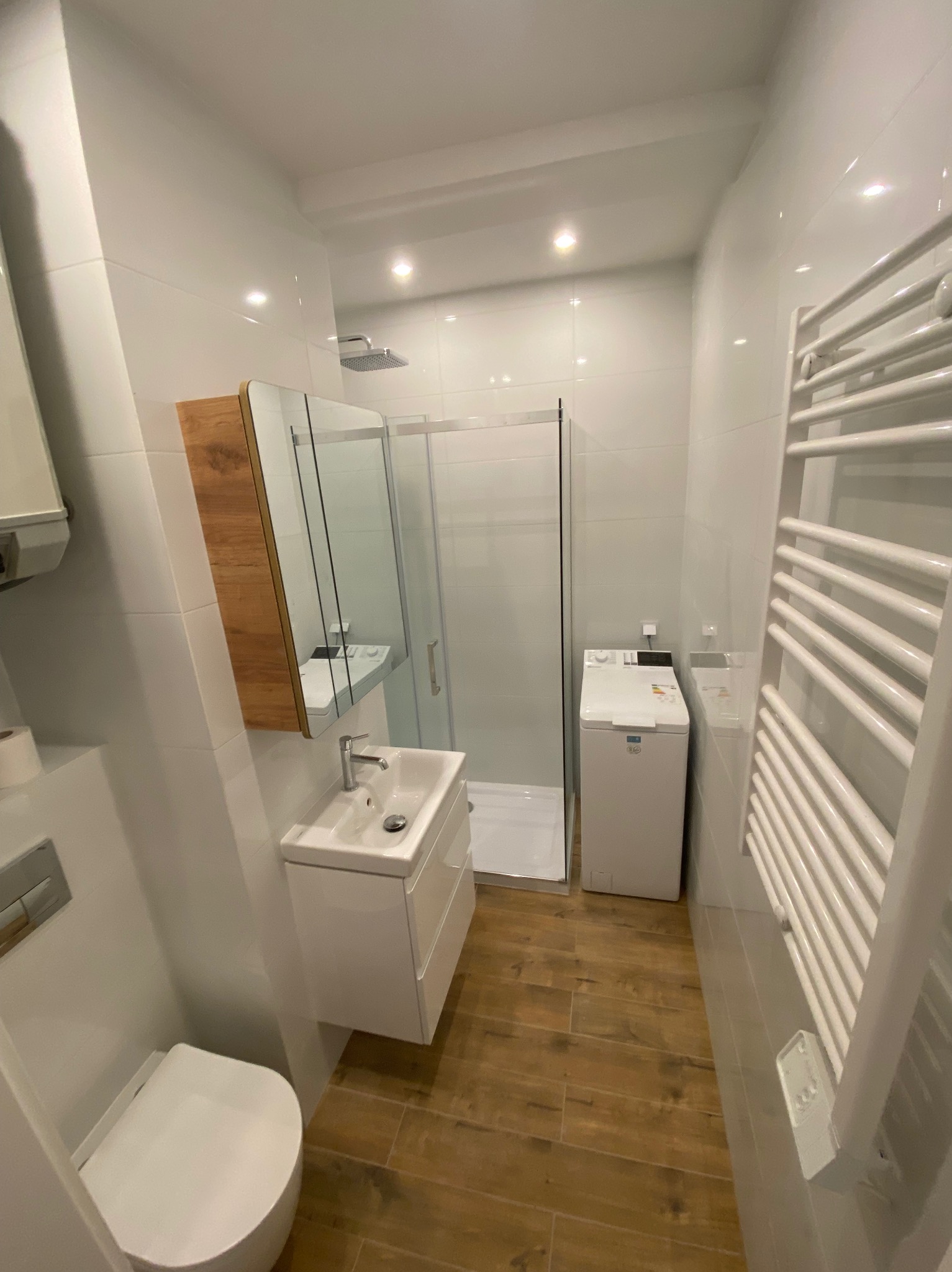Jasna łazienka z kabiną prysznicową, umywalką z szafką, toaletą i grzejnikiem drabinkowym. Podłoga wyłożona panelami imitującymi drewno. Widok z perspektywy.