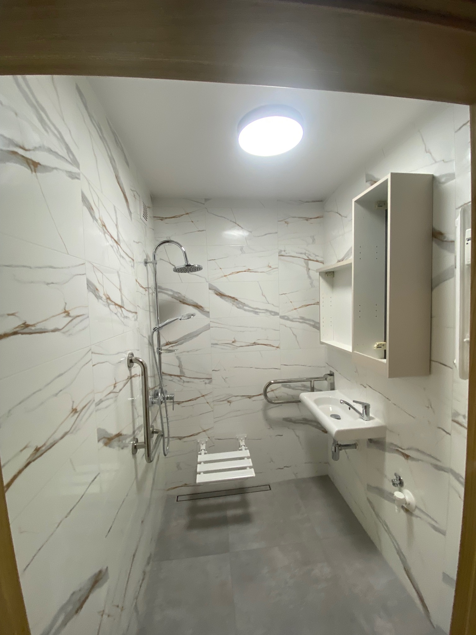 Nowoczesna łazienka z białymi płytkami imitującymi marmur, szarą podłogą, kabiną prysznicową bez brodzika i udogodnieniami dla osób z niepełnosprawnościami.