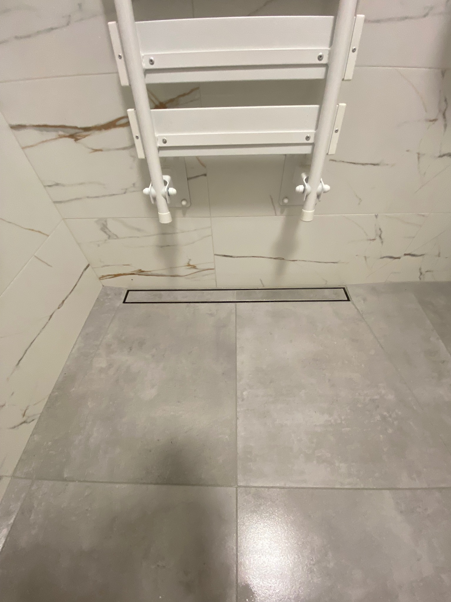 Nowoczesna łazienka z szarymi płytkami podłogowymi, odpływem liniowym i białym grzejnikiem drabinkowym na ścianie z marmurowymi płytkami. Minimalistyczny design.