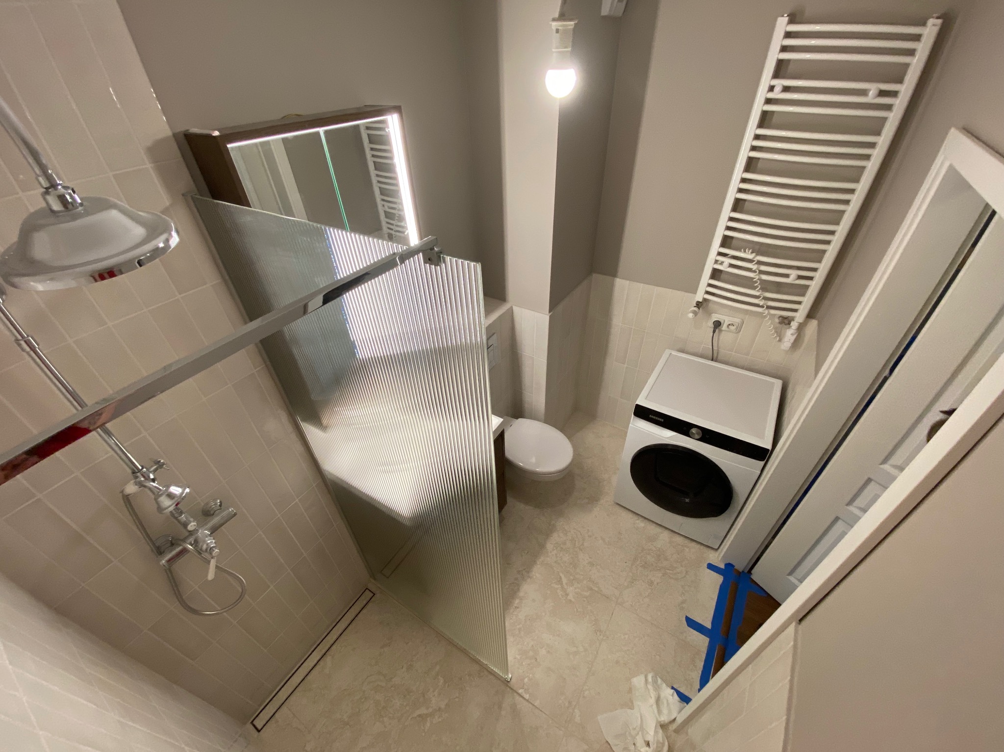 Nowoczesna łazienka po remoncie: kabina prysznicowa ze szklaną ścianką, grzejnik drabinkowy, pralka i toaleta w jasnej tonacji. Widok z góry.