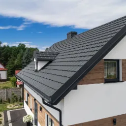 Nowoczesny dom jednorodzinny z ciemnoszarą dachówką modułową, czarnymi rynnami i obróbkami blacharskimi, widok z góry na tle błękitnego nieba i zieleni drzew.