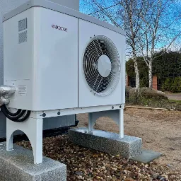 Biała, zewnętrzna jednostka pompy ciepła Stiebel Eltron, zamontowana na granitowych blokach, z widocznymi rurami i wentylatorem, na tle ogrodu z brzozami.