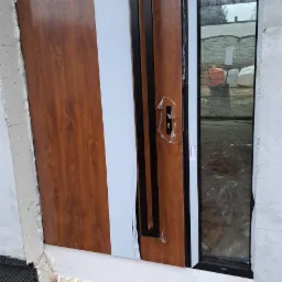 Brązowe drzwi wejściowe z długim czarnym uchwytem, częściowo zabezpieczone folią ochronną, osadzone w surowym otworze ściennym z widoczną pianką montażową i fragmentami liści na ziemi.