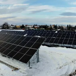 Rozległa instalacja paneli słonecznych na zasypanym śniegiem terenie, widoczne zabudowania w tle, drabina oparta o jeden z paneli.