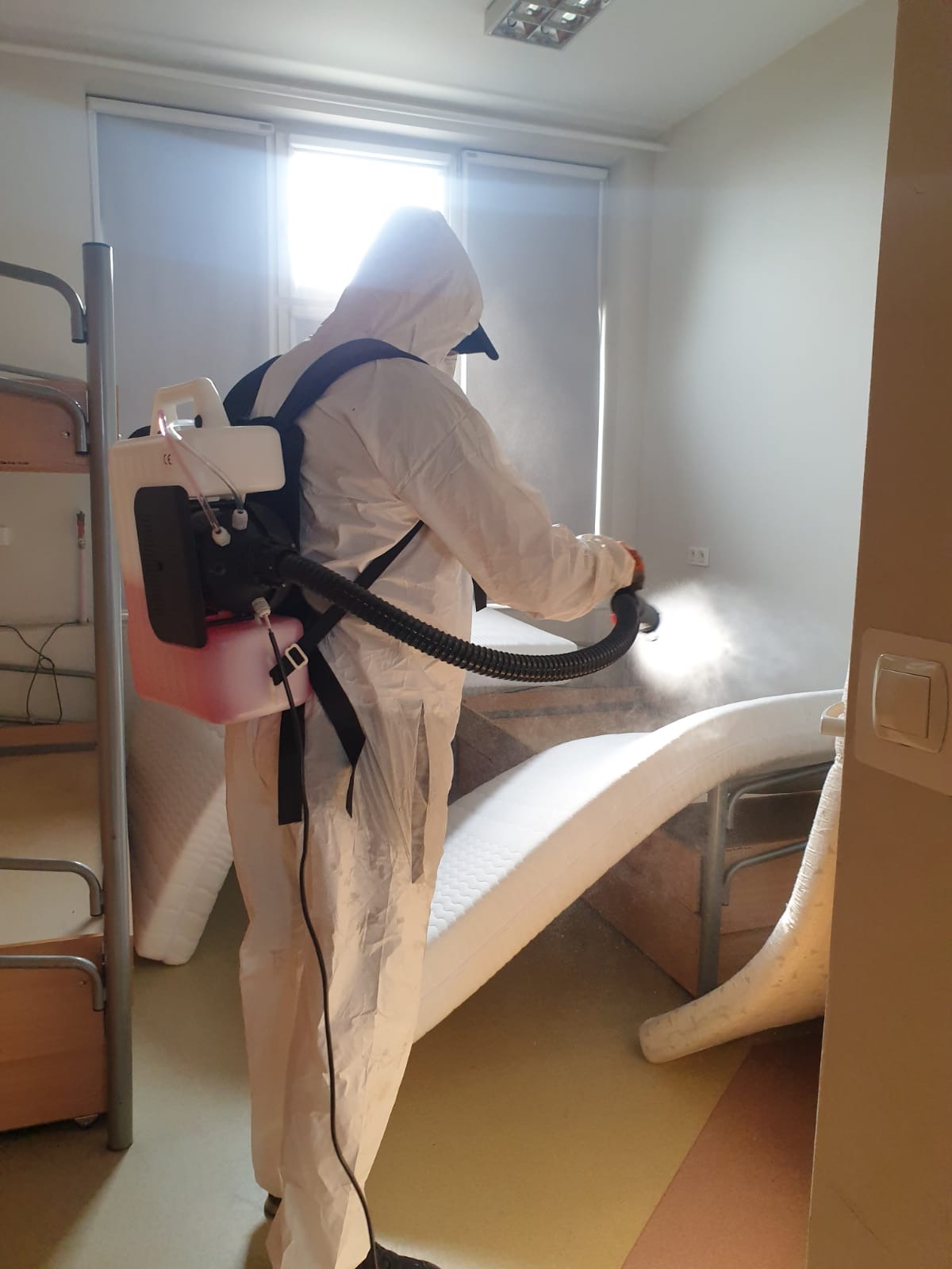 Osoba w kombinezonie ochronnym z plecakowym opryskiwaczem, rozpyla środek na łóżko piętrowe w jasnym pomieszczeniu.