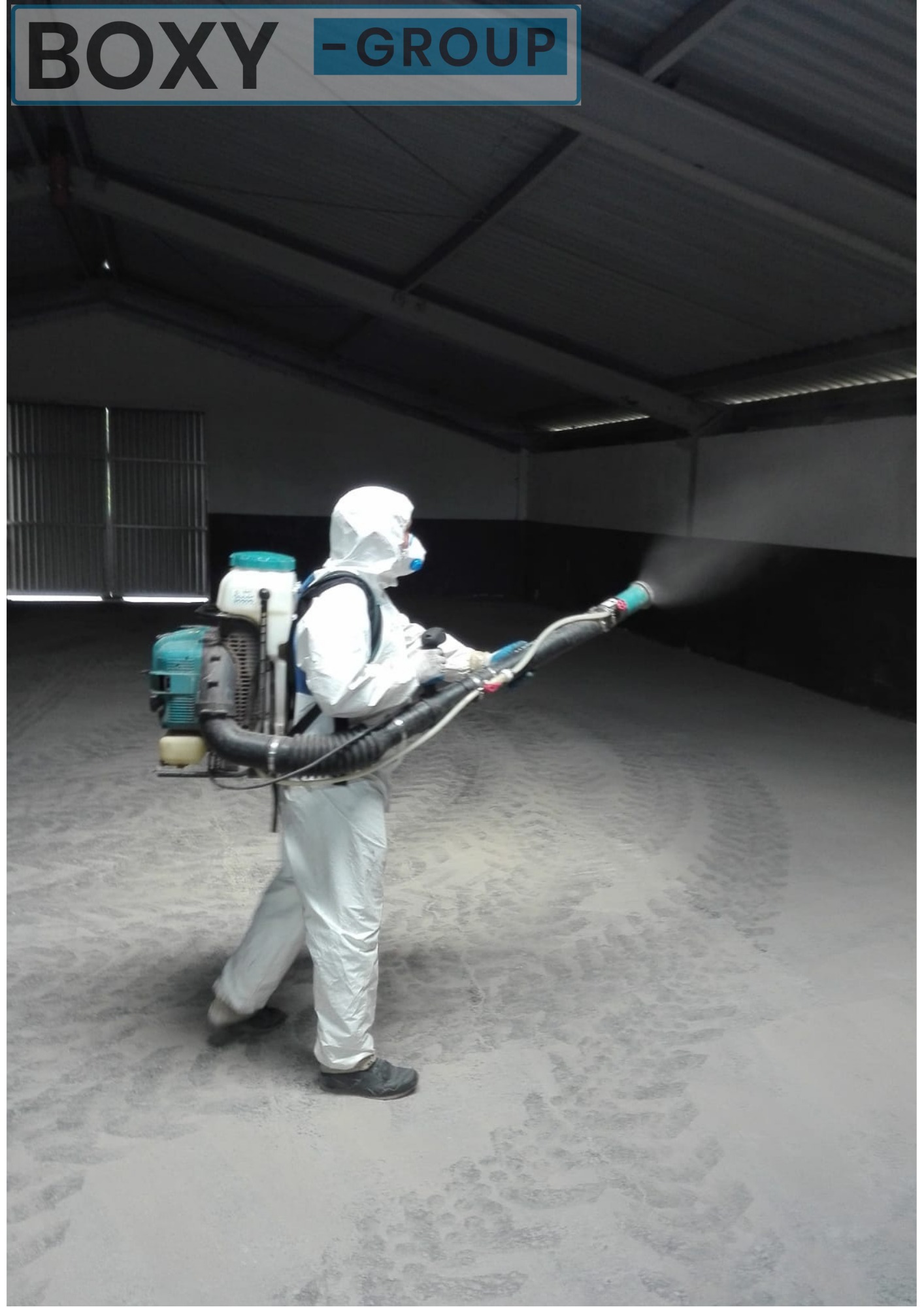 Pracownik w kombinezonie ochronnym i masce rozpyla środek dezynfekujący wewnątrz magazynu z betonową podłogą, widoczne ślady opon na podłodze.