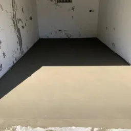 Świeżo wylana, gładka posadzka jastrychowa w surowym, odrapanym pomieszczeniu z małym okienkiem kratowanym i lampą na ścianie, widoczny cień osoby robiącej zdjęcie.