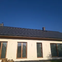 Dom jednorodzinny z ocieploną elewacją w kolorze kremowym, z ciemnoszarym dachem z dachówki ceramicznej i dwoma kominami; okna zabezpieczone folią ochronną, widoczne ślady po montażu na elewacji.