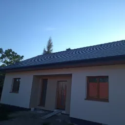 Jasny dom jednorodzinny z nową, grafitową dachówką i oknami w drewnianych ramach, świeżo po nałożeniu tynku zewnętrznego, widoczne niedokończone prace wykończeniowe wokół budynku.