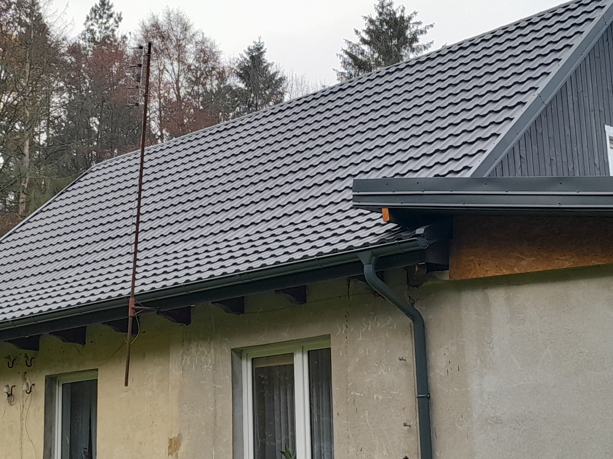 Dom z nowym, grafitowym dachem z blachodachówki, widoczne rynny i antena na tle drzew.