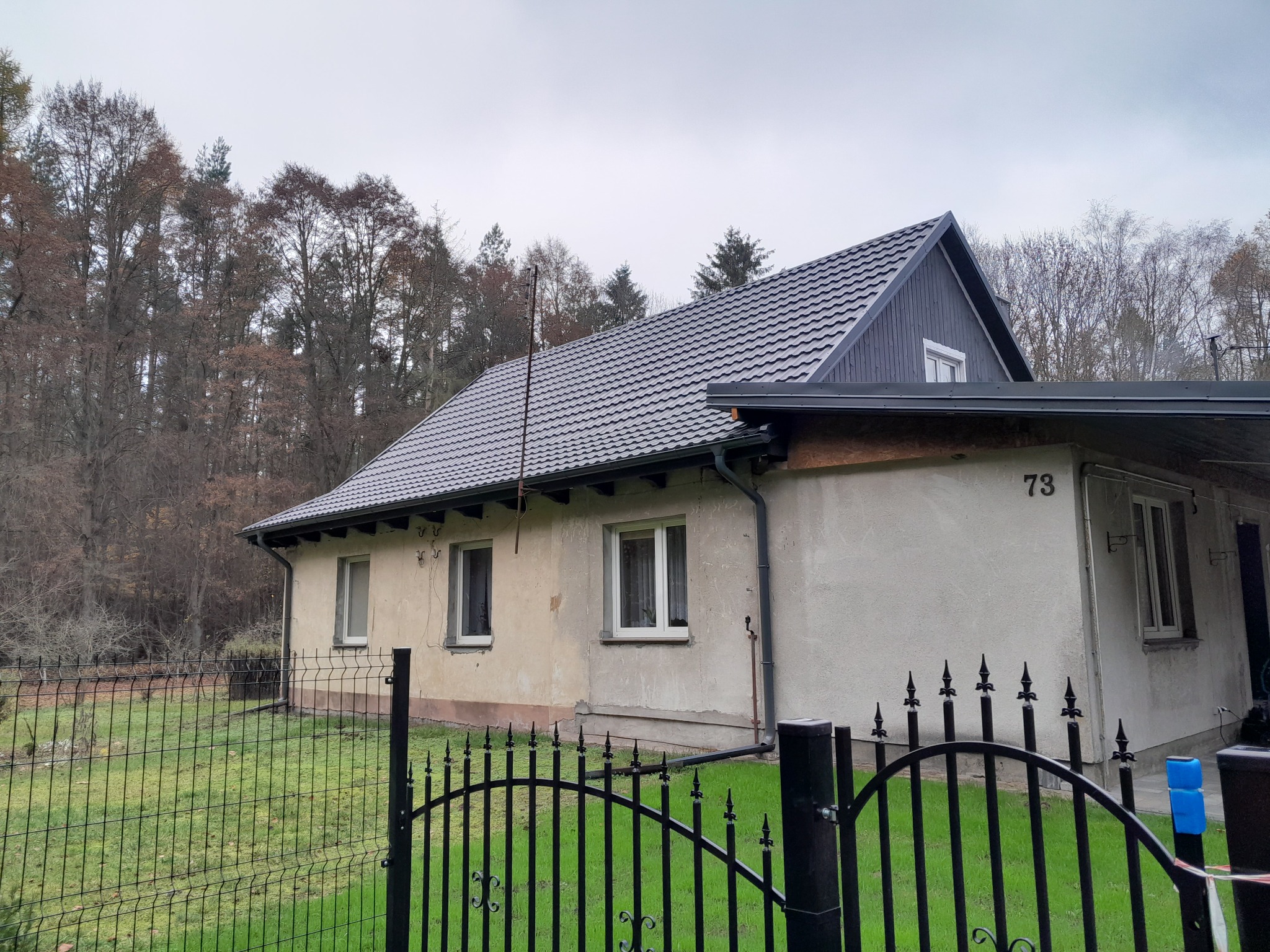 Niski dom jednorodzinny z szarą dachówką i fragmentami nieocieplonej elewacji, widoczny przez czarne, metalowe ogrodzenie na tle drzew bez liści.