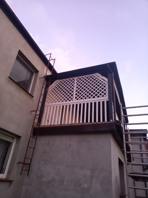 Elewacja budynku z balkonem z białą, ażurową balustradą, widok z dołu, z przystawioną metalową drabiną. Ściana budynku szara, tynkowana.
