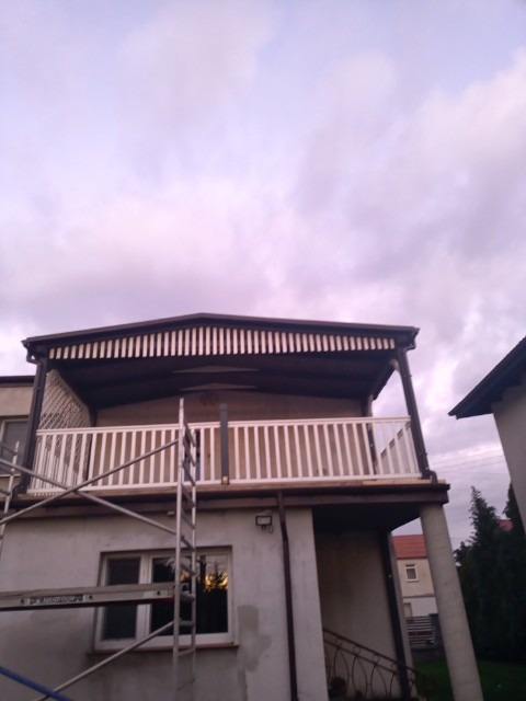 Elewacja domu z balkonem w trakcie renowacji, widoczne rusztowanie i odnowiona balustrada balkonu na tle pochmurnego nieba.