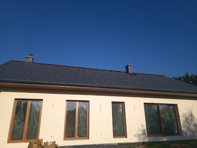 Dom jednorodzinny z ocieploną elewacją w kolorze kremowym, z ciemnoszarym dachem z dachówki ceramicznej i dwoma kominami; okna zabezpieczone folią ochronną, widoczne ślady po montażu na elewacji.