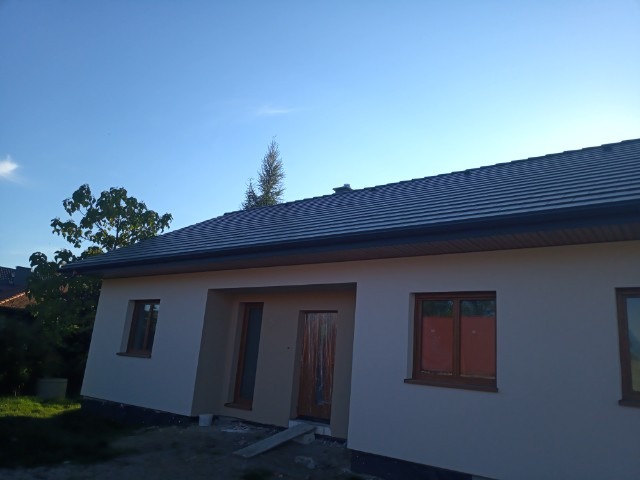 Jasny dom jednorodzinny z nową, grafitową dachówką i oknami w drewnianych ramach, świeżo po nałożeniu tynku zewnętrznego, widoczne niedokończone prace wykończeniowe wokół budynku.