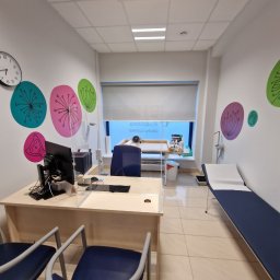 ReklaMaster MONIKA SZCZĘSNA - Wnętrze gabinetu lekarskiego z biurkiem oddzielonym szybą ochronną, leżanką i dekoracyjnymi naklejkami na ścianie w kształcie kwiatów i sowy.