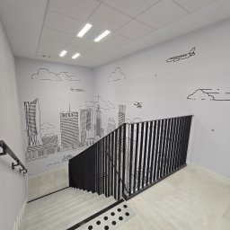 ReklaMaster MONIKA SZCZĘSNA - Wnętrze klatki schodowej z minimalistycznym rysunkiem panoramy miasta na ścianie, obejmującym wieżowce, samolot i helikopter w stylu line art.