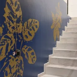 Klatka schodowa z granatową ścianą ozdobioną dużymi, złotymi motywami roślinnymi, widoczne schody wyłożone jasnymi płytkami imitującymi drewno.
