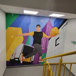 Ilustracja na ścianie w klatce schodowej przedstawiająca kuriera z paczką na tle miasta i firmowego samochodu dostawczego.