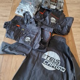 ReklaMaster MONIKA SZCZĘSNA - Szare ubrania robocze z nadrukowanym logo firmy TELS CARGO, niektóre w oryginalnych foliowych opakowaniach z napisem FOR ECO, leżące na drewnianym stole.