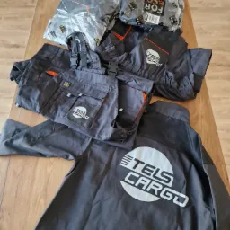 Szare ubrania robocze z nadrukowanym logo firmy TELS CARGO, niektóre w oryginalnych foliowych opakowaniach z napisem FOR ECO, leżące na drewnianym stole.