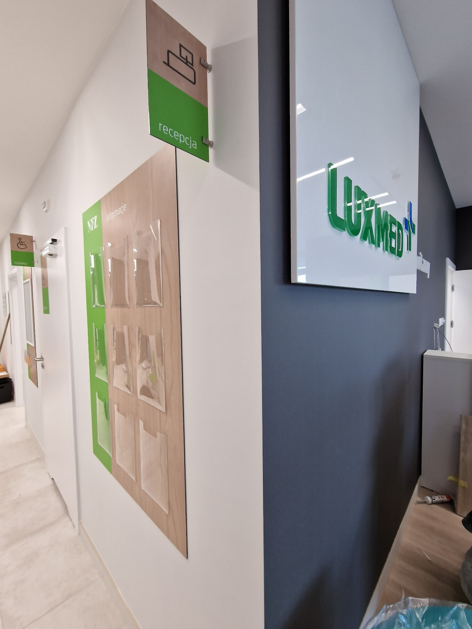 Tabliczki informacyjne w placówce medycznej: oznaczenie recepcji, logo Luxmed na ścianie i tablica na ulotki NFZ.
