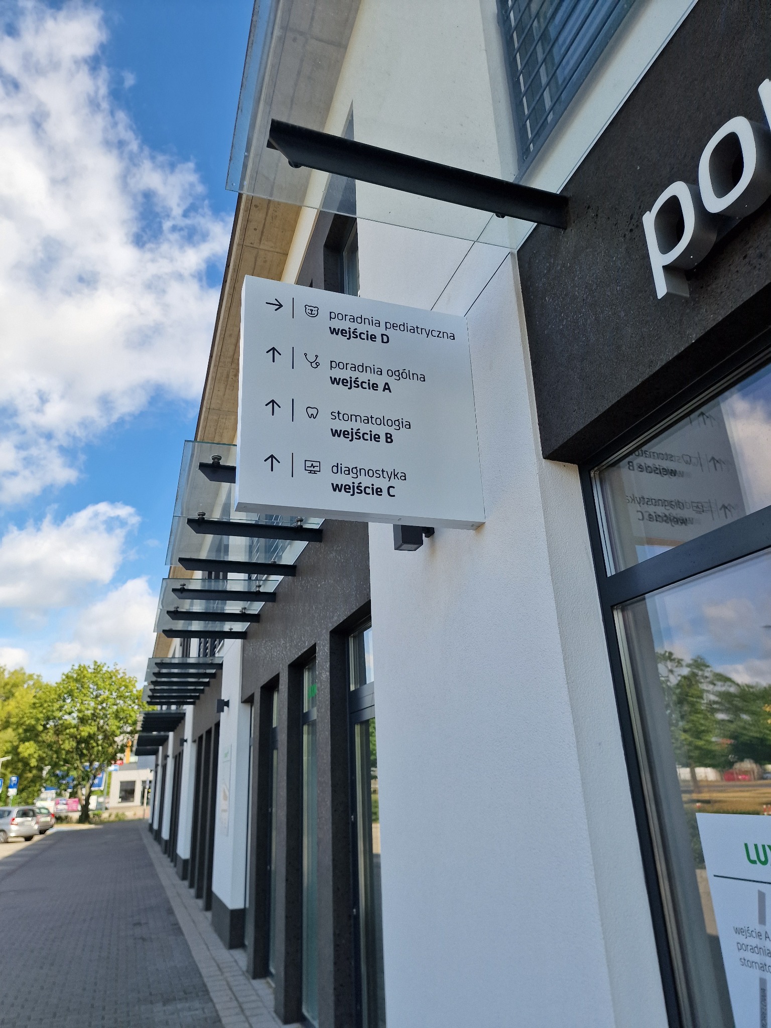 Tablica informacyjna przy wejściu do budynku z listą poradni: pediatryczna, ogólna, stomatologia, diagnostyka i odpowiadającymi im wejściami A, B, C, D.