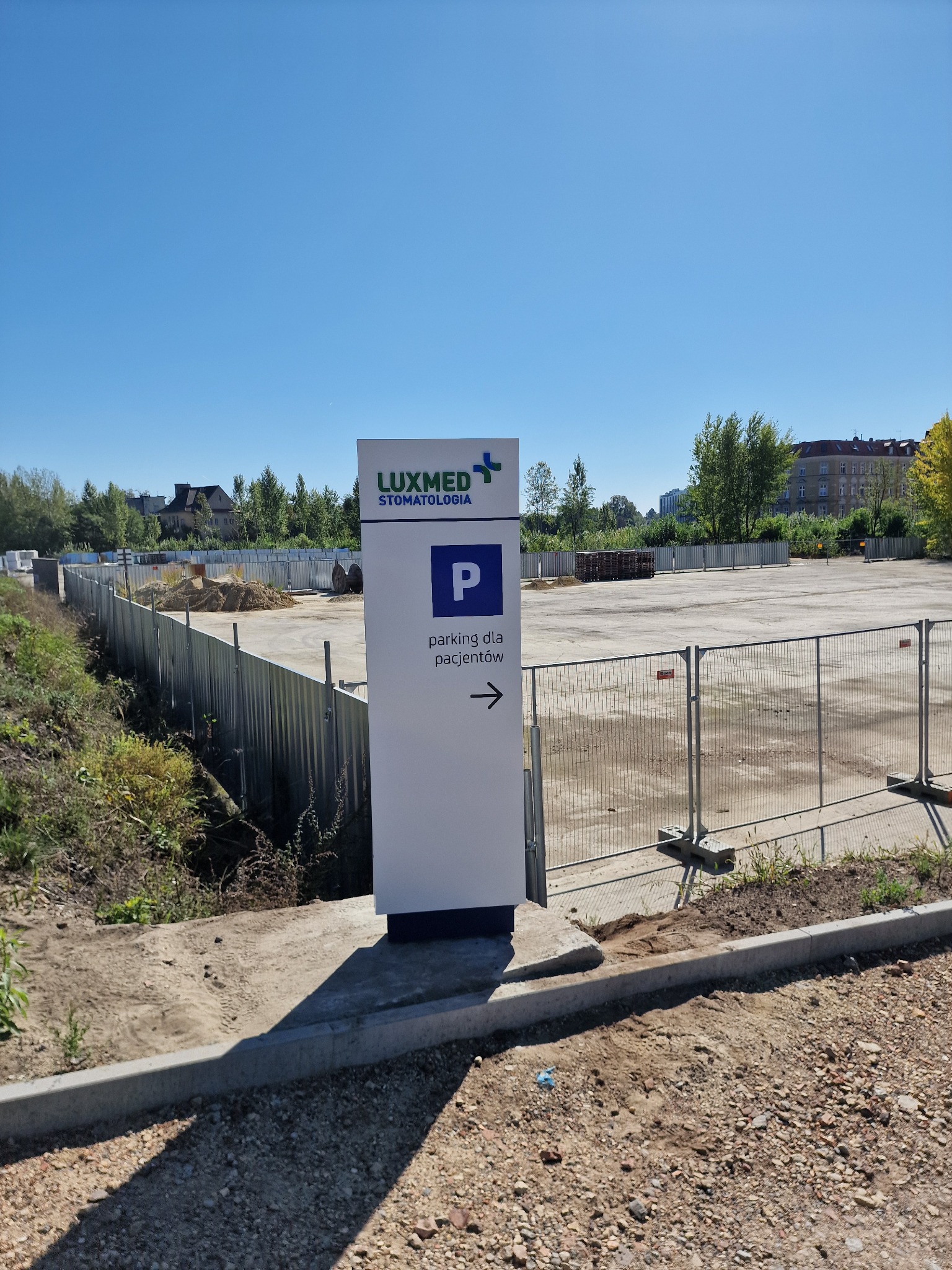 Tablica informacyjna Luxmed Stomatologia z symbolem parkingu dla pacjentów, umieszczona na tle placu budowy z widocznymi ogrodzeniami i składowanym materiałem.