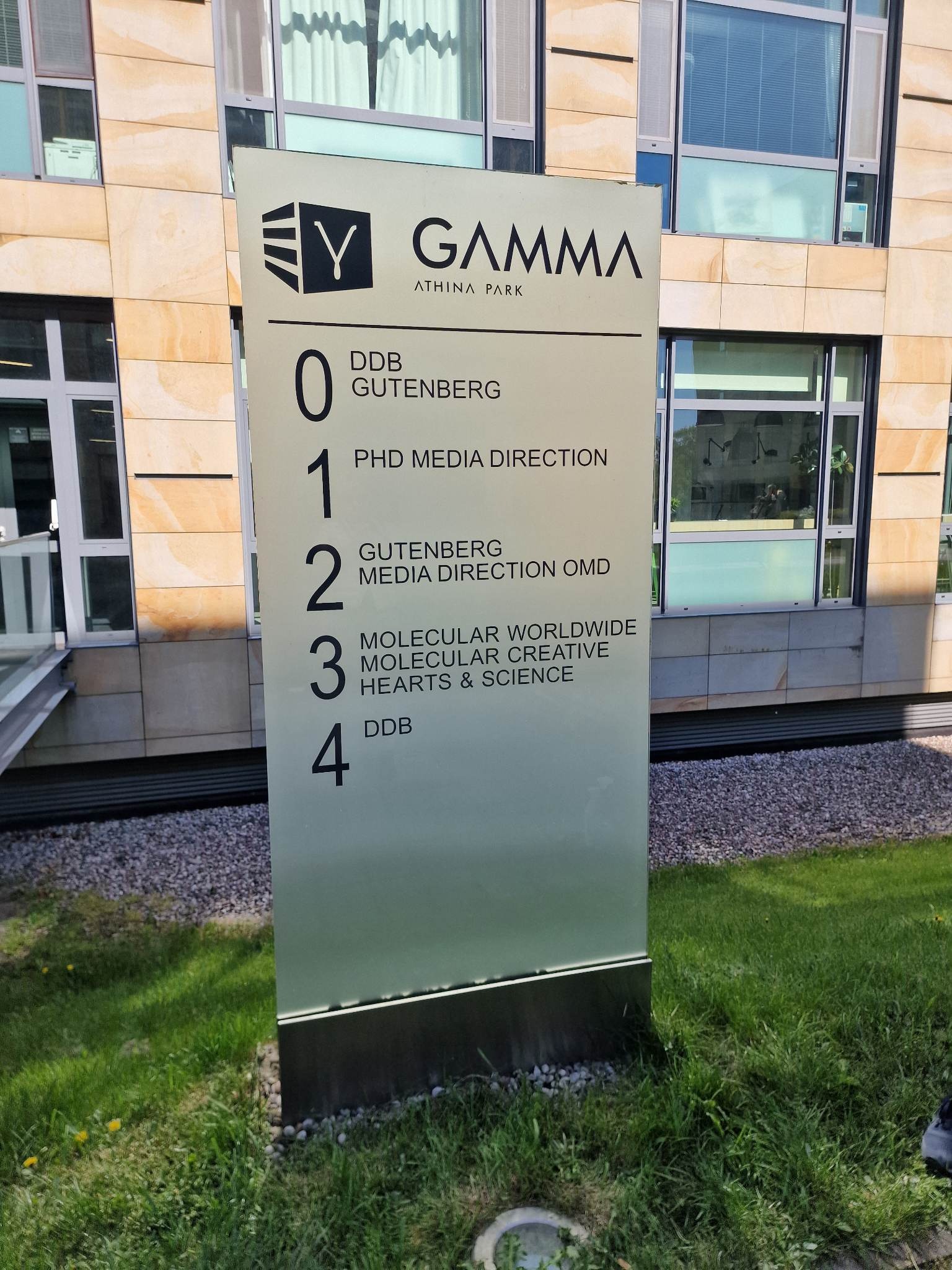 Pylon informacyjny z logo Gamma Athina Park i listą firm w budynku, na tle elewacji budynku z kamienia.