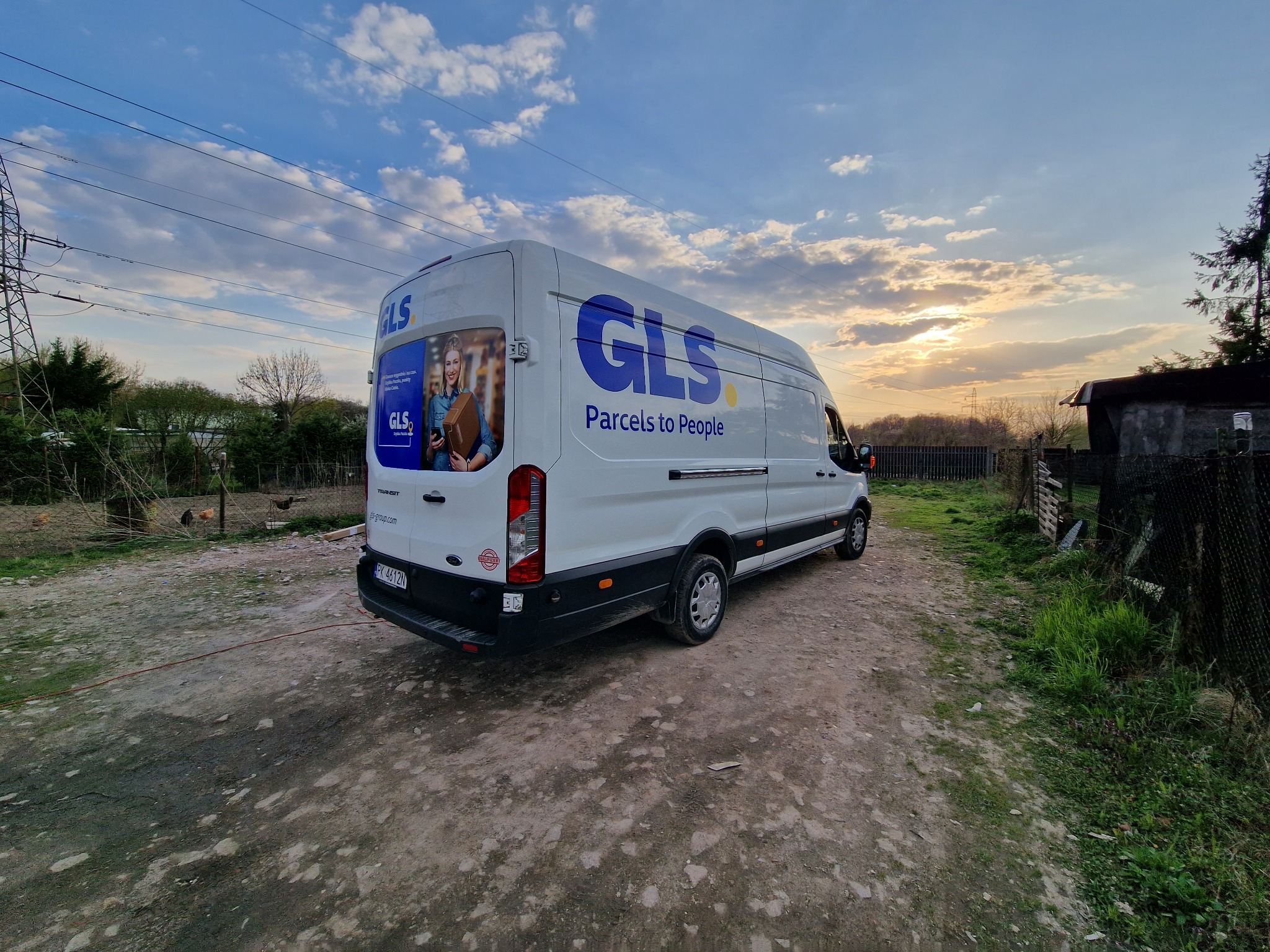 Biały samochód dostawczy GLS z nadrukowanym logo i wizerunkiem osoby, stojący na polnej drodze o zmierzchu, w tle słupy wysokiego napięcia i zabudowania gospodarcze.