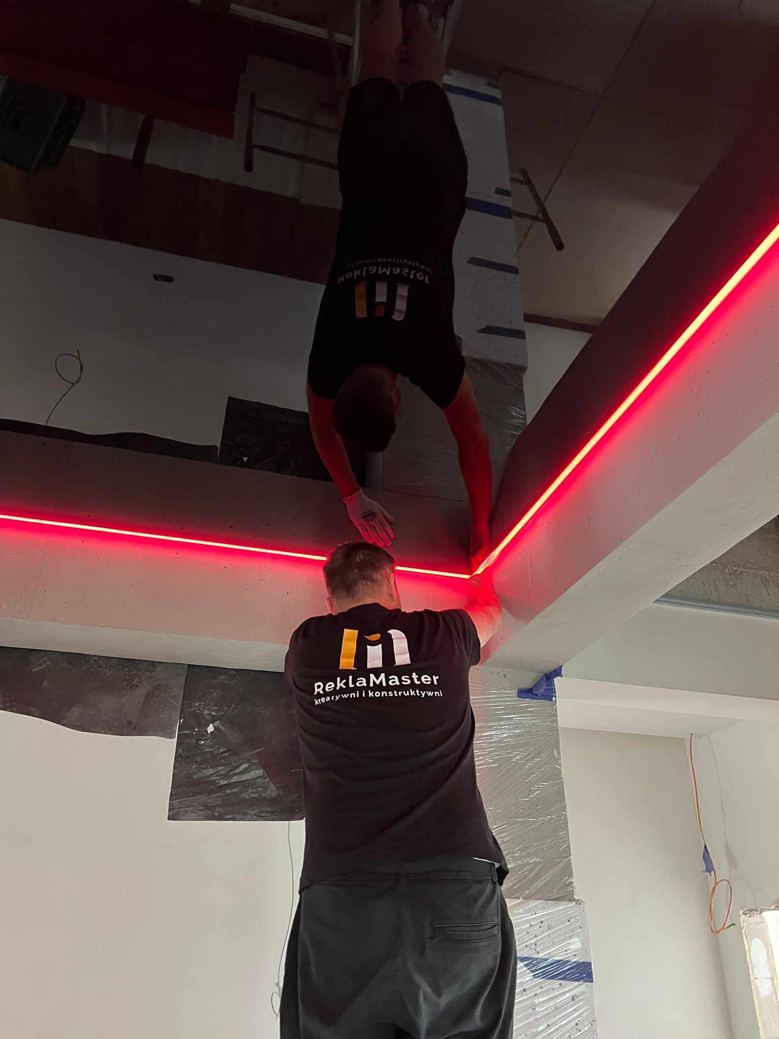 Dwóch pracowników montuje czerwoną listwę LED na konstrukcji sufitowej, jeden z nich w rękawicach, obaj w koszulkach z logo firmy.