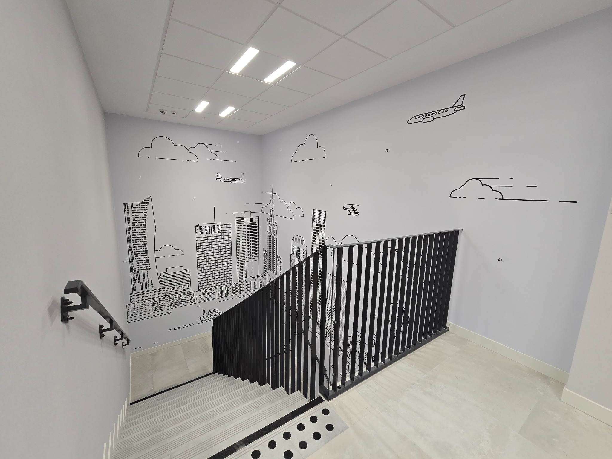 Wnętrze klatki schodowej z minimalistycznym rysunkiem panoramy miasta na ścianie, obejmującym wieżowce, samolot i helikopter w stylu line art.