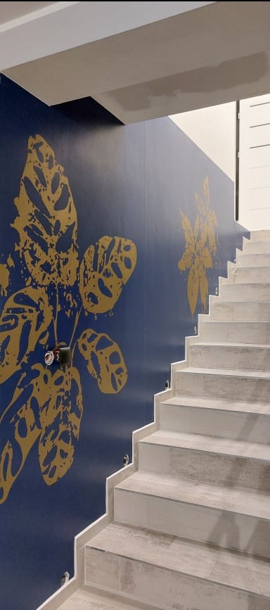Klatka schodowa z granatową ścianą ozdobioną dużymi, złotymi motywami roślinnymi, widoczne schody wyłożone jasnymi płytkami imitującymi drewno.