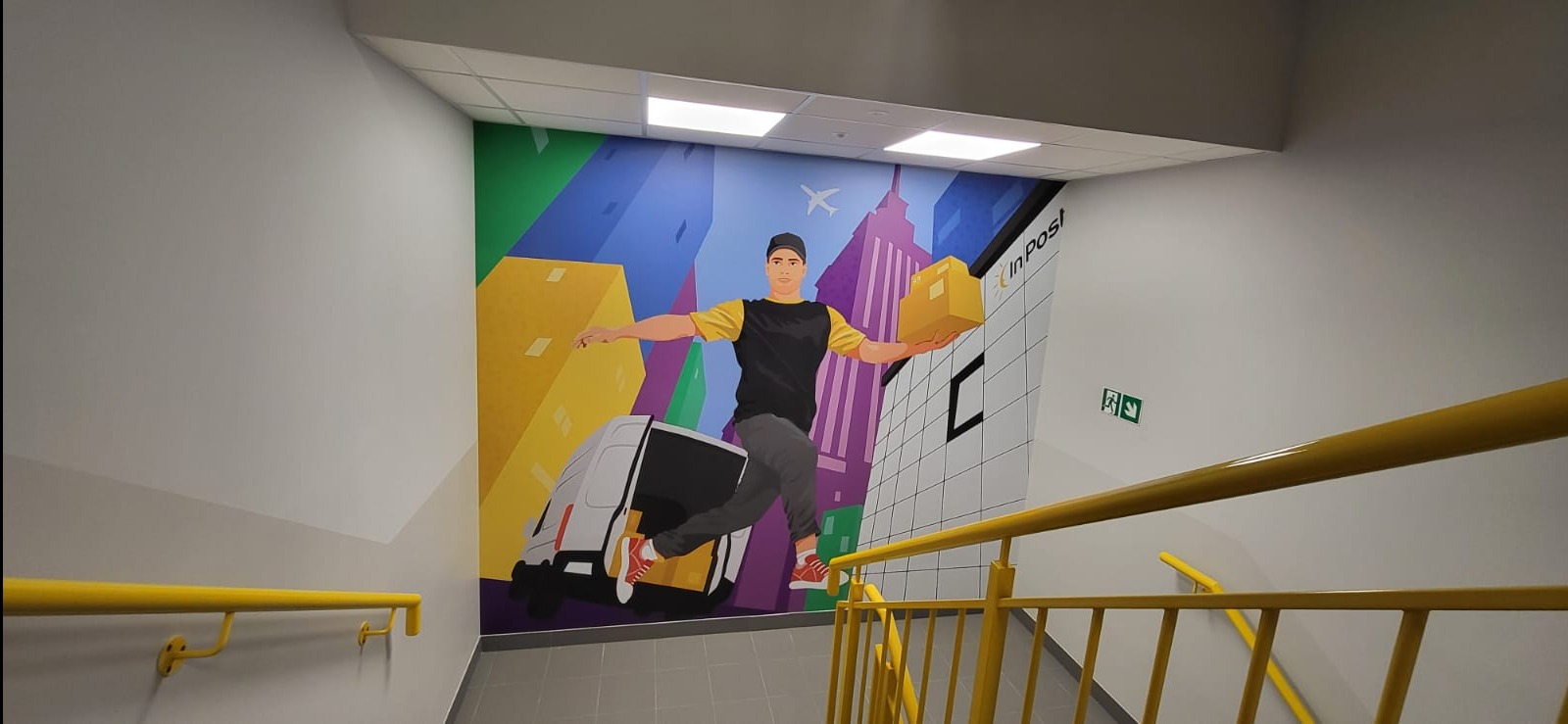 Ilustracja na ścianie w klatce schodowej przedstawiająca kuriera z paczką na tle miasta i firmowego samochodu dostawczego.