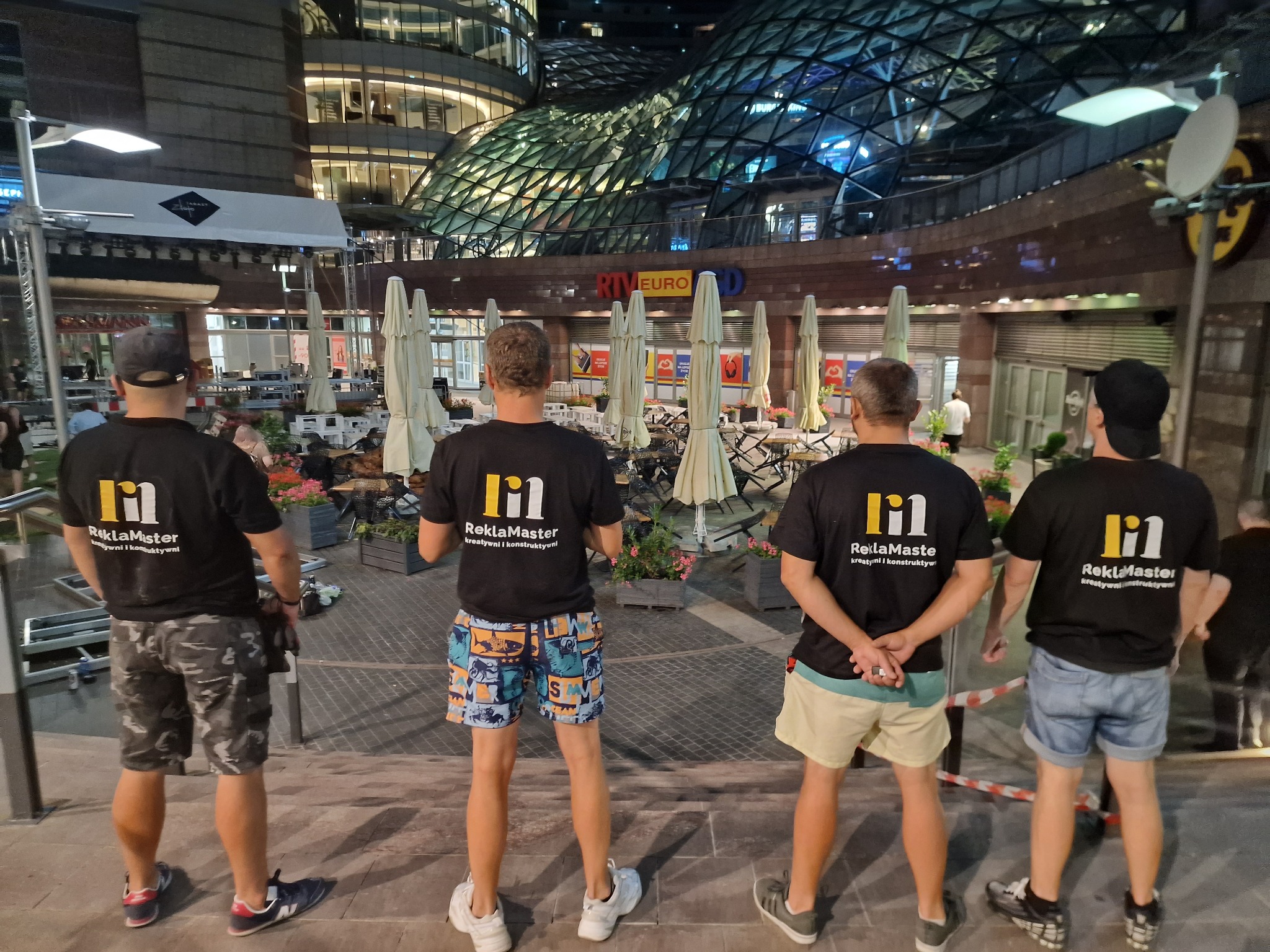 Czterech mężczyzn w czarnych koszulkach z logo firmy 'ReklaMaster' stoi tyłem na schodach na tle budynku z przeszkloną fasadą i restauracji z parasolami.