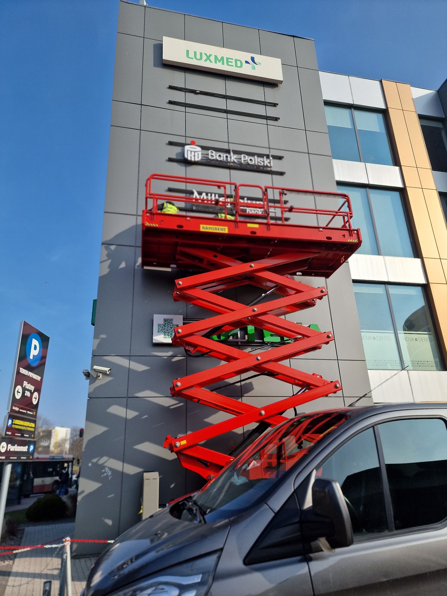Montaż szyldu Luxmed i Bank Polski na elewacji budynku biurowego z użyciem podnośnika koszowego, widoczna część zaparkowanego samochodu i szyld informacyjny parkingu.