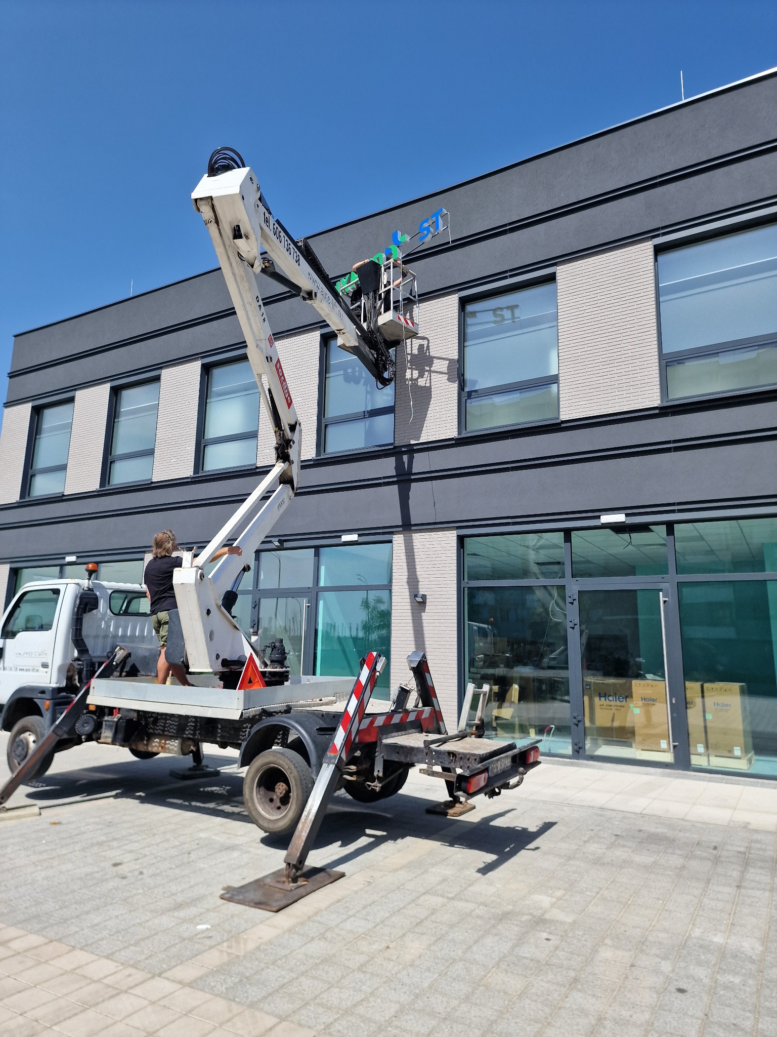 Montaż niebieskiego napisu na elewacji budynku z użyciem podnośnika koszowego, widoczny fragment napisu 'LST', pracownik w koszu podnośnika, drugi pracownik na platformie podnośnika.