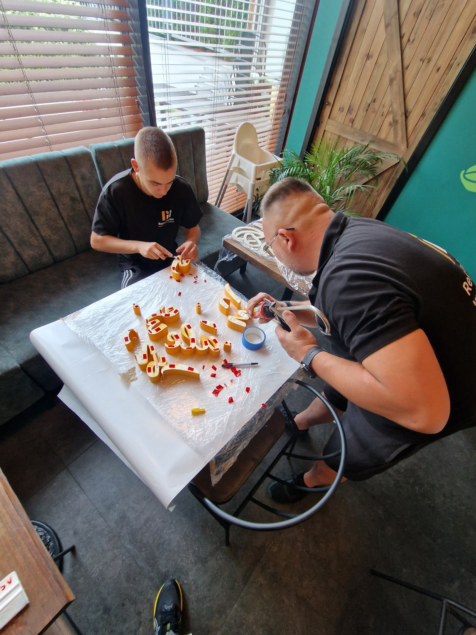 Dwóch mężczyzn przy stole tworzy napisy z żółtych elementów i czerwonych akcentów, używając narzędzi do precyzyjnego montażu. Na stole folia ochronna i drobne pozostałości materiałów.