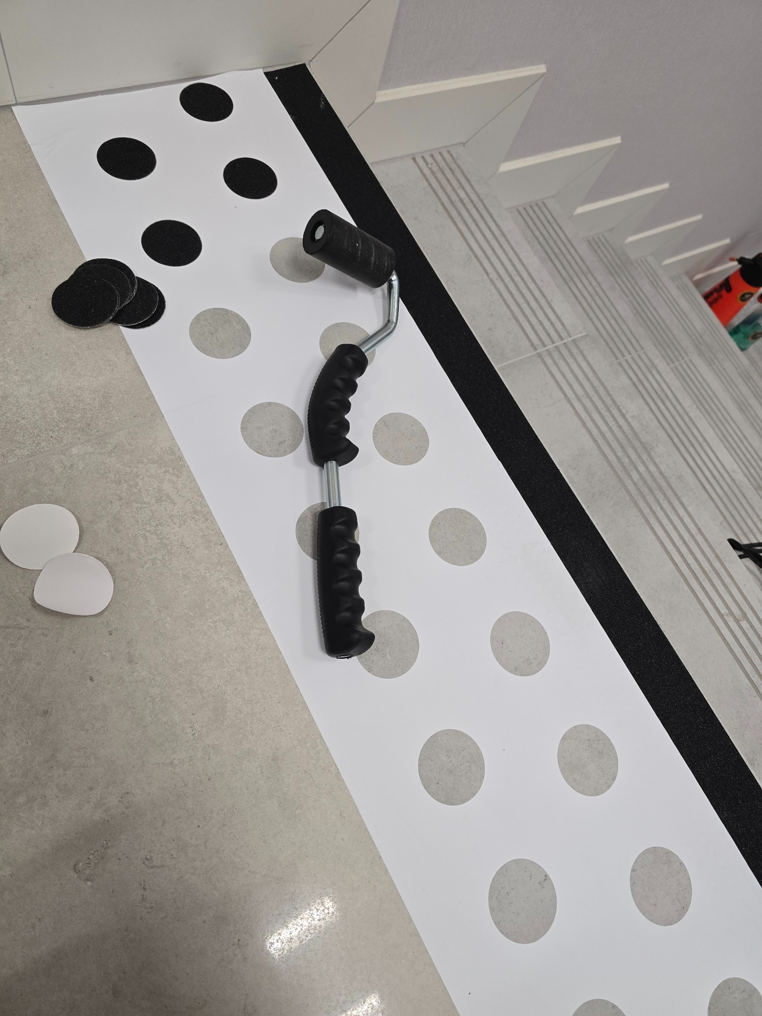 Aplikacja naklejek podłogowych z motywem kropek, wałek do naklejek i odklejone okrągłe elementy na szarej podłodze w pobliżu schodów.
