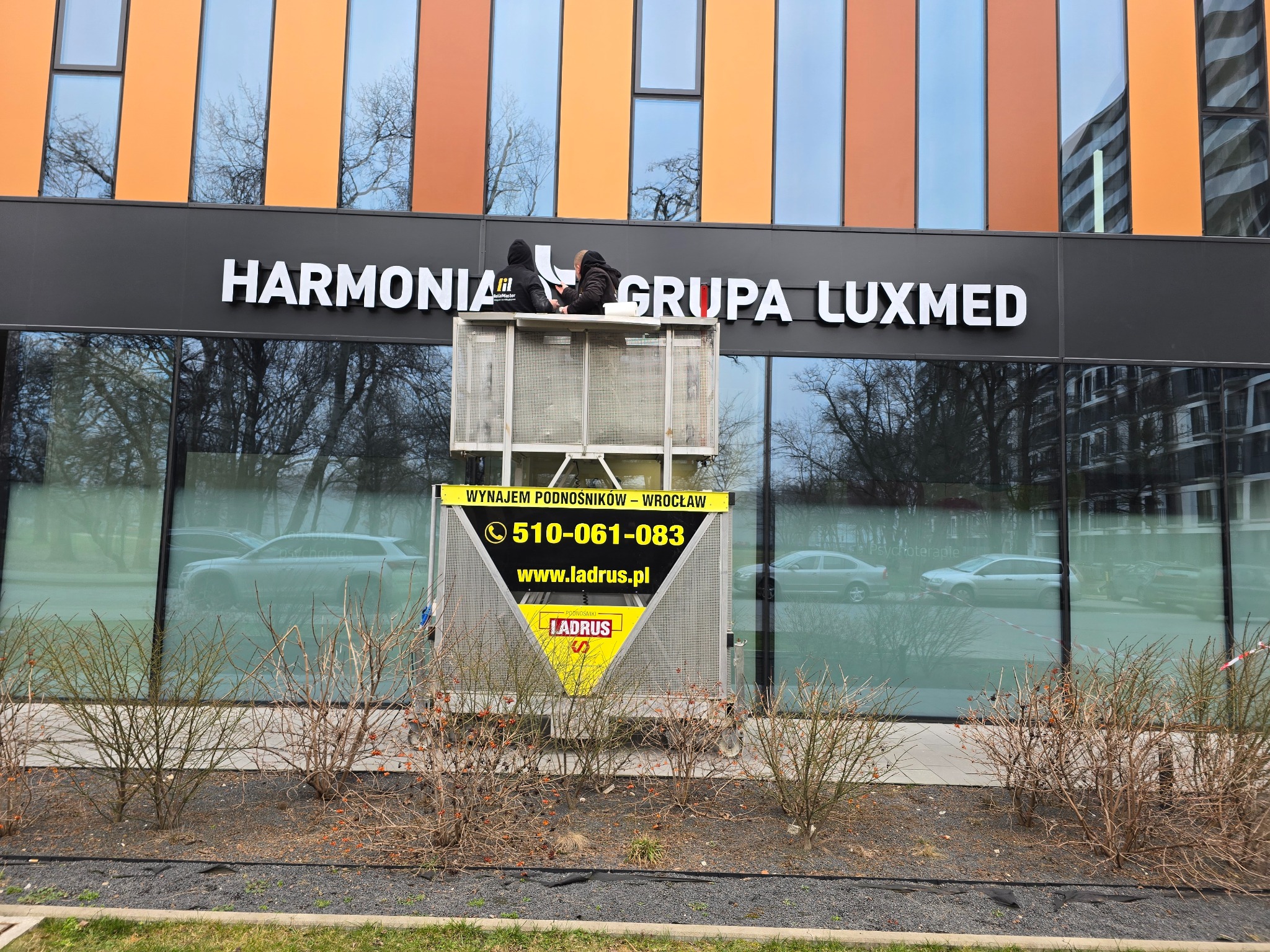 Pracownicy na podnośniku montują podświetlany szyld z nazwą Harmonia Grupa Luxmed na elewacji budynku z pomarańczowo-niebieskimi panelami.