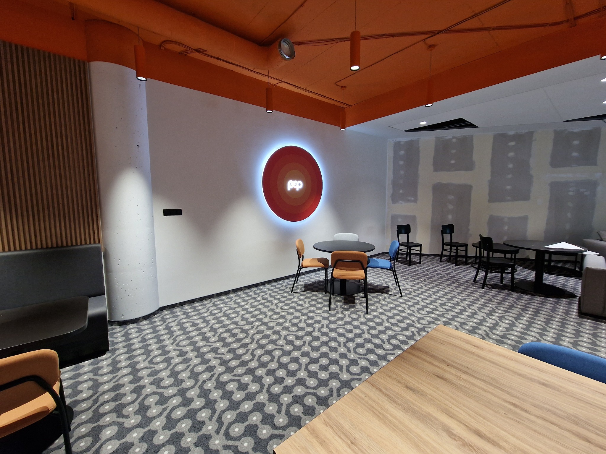Wnętrze biura z pomarańczowym sufitem, wzorzystą wykładziną, okrągłym podświetlanym logo na ścianie, stołami i krzesłami w różnych kolorach.