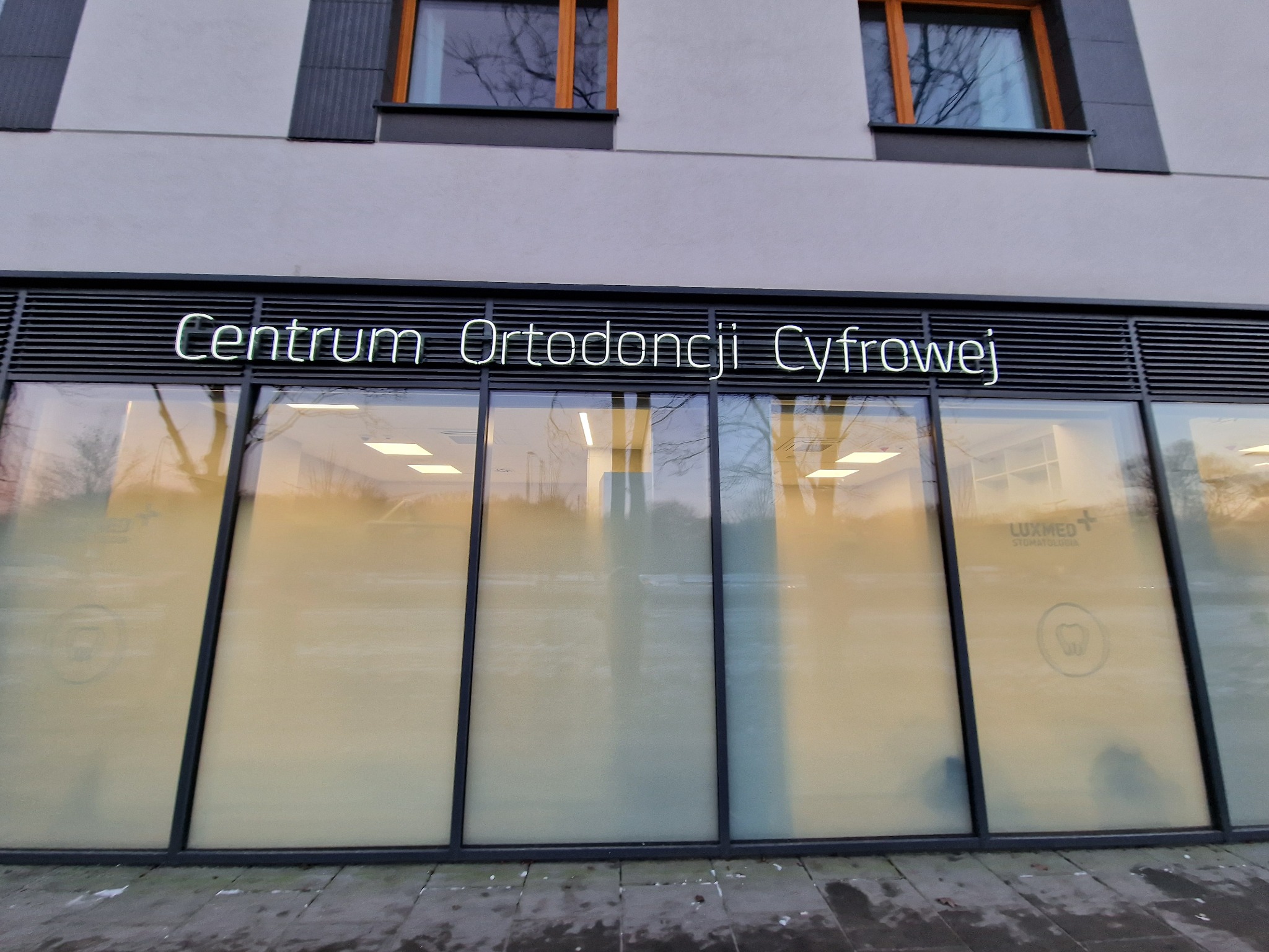 Elewacja budynku z dużymi oknami, nad którymi umieszczono podświetlany napis 'Centrum Ortodoncji Cyfrowej'. Na szybach okien widoczne logo Luxmed Stomatologia i grafika zęba.