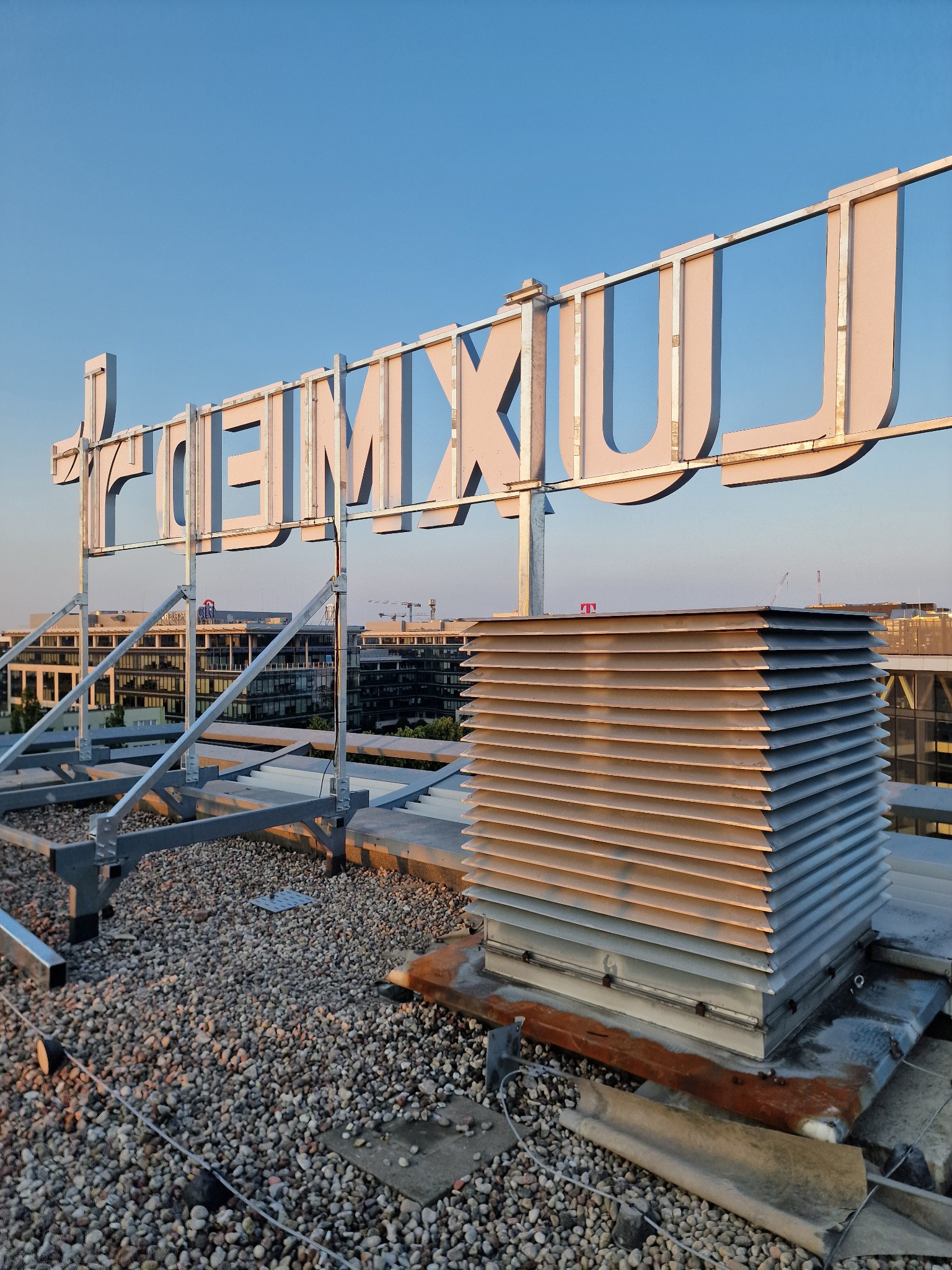 Duże, przestrzenne litery tworzące napis 'PREMIUM U' na dachu budynku, widoczne z bliska, z elementami konstrukcyjnymi i wentylacją, na tle panoramy miasta w słoneczny dzień.