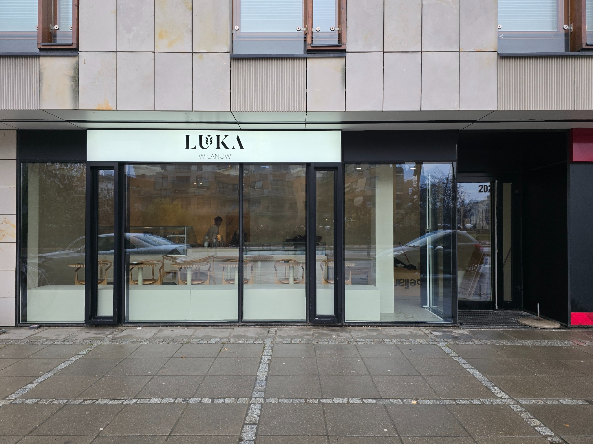 Fasada lokalu usługowego z dużymi oknami wystawowymi i szyldem 'LUKA Wilanów' nad wejściem, widok z zewnątrz na chodnik i ulicę.