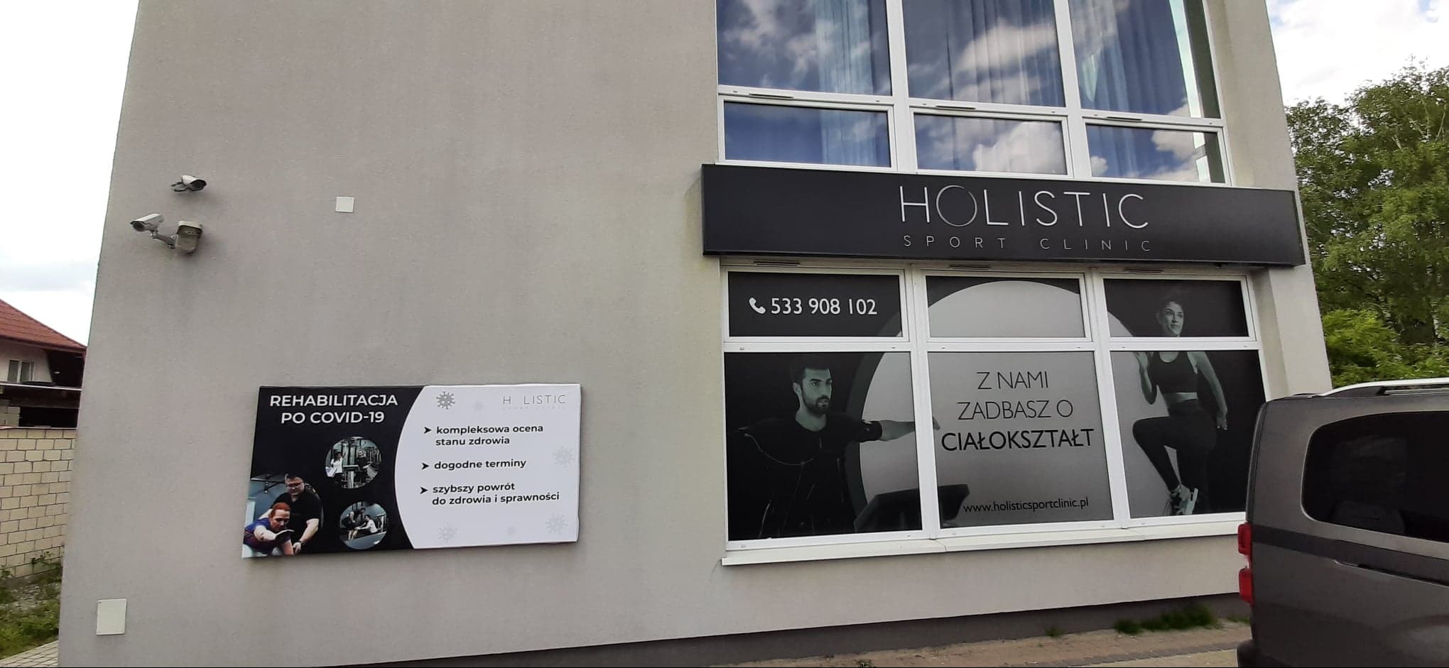 Zewnętrzny widok budynku Holistic Sport Clinic z reklamami na ścianie i oknach, w tym tablica informująca o rehabilitacji po COVID-19 i folie okienne z grafikami osób ćwiczących i numerem telefonu.