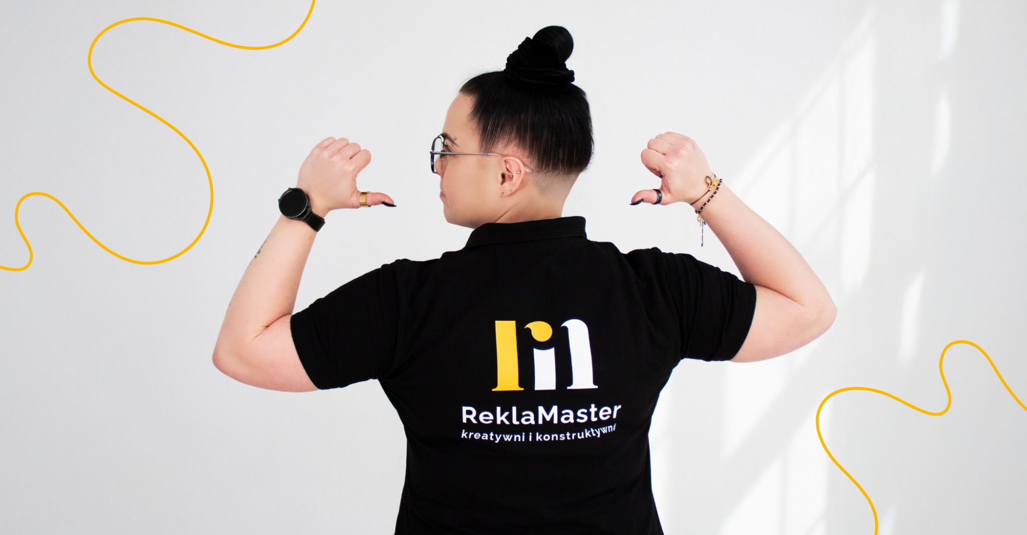 Osoba w czarnej koszulce polo z logo ReklaMaster na plecach, prezentująca firmę. Widok z tyłu, na białym tle z żółtymi elementami graficznymi.