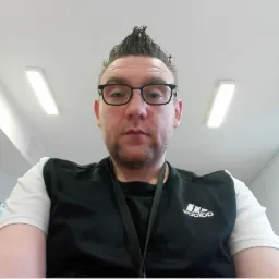 Portret mężczyzny w okularach z irokezem, ubrany w sportową koszulkę z logo Adidas, na tle białego sufitu z lampami.