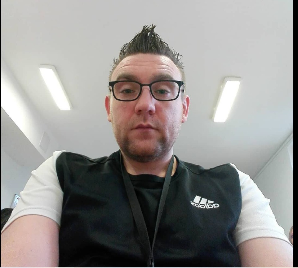 Portret mężczyzny w okularach z irokezem, ubrany w sportową koszulkę z logo Adidas, na tle białego sufitu z lampami.